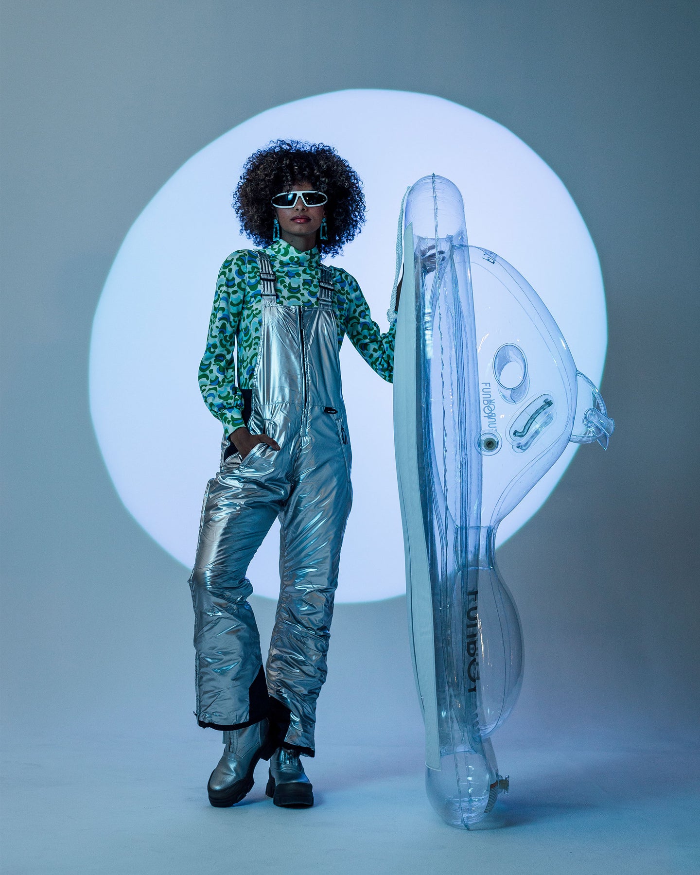 Women is standing holding up the Funboy Super Clear Snowmobile Snow Sled. She's posing in a metallic silver ski suit with futuristic sunglasses & metallic silver boots. The background is grey with a white/blue colored circled that's lit up. 