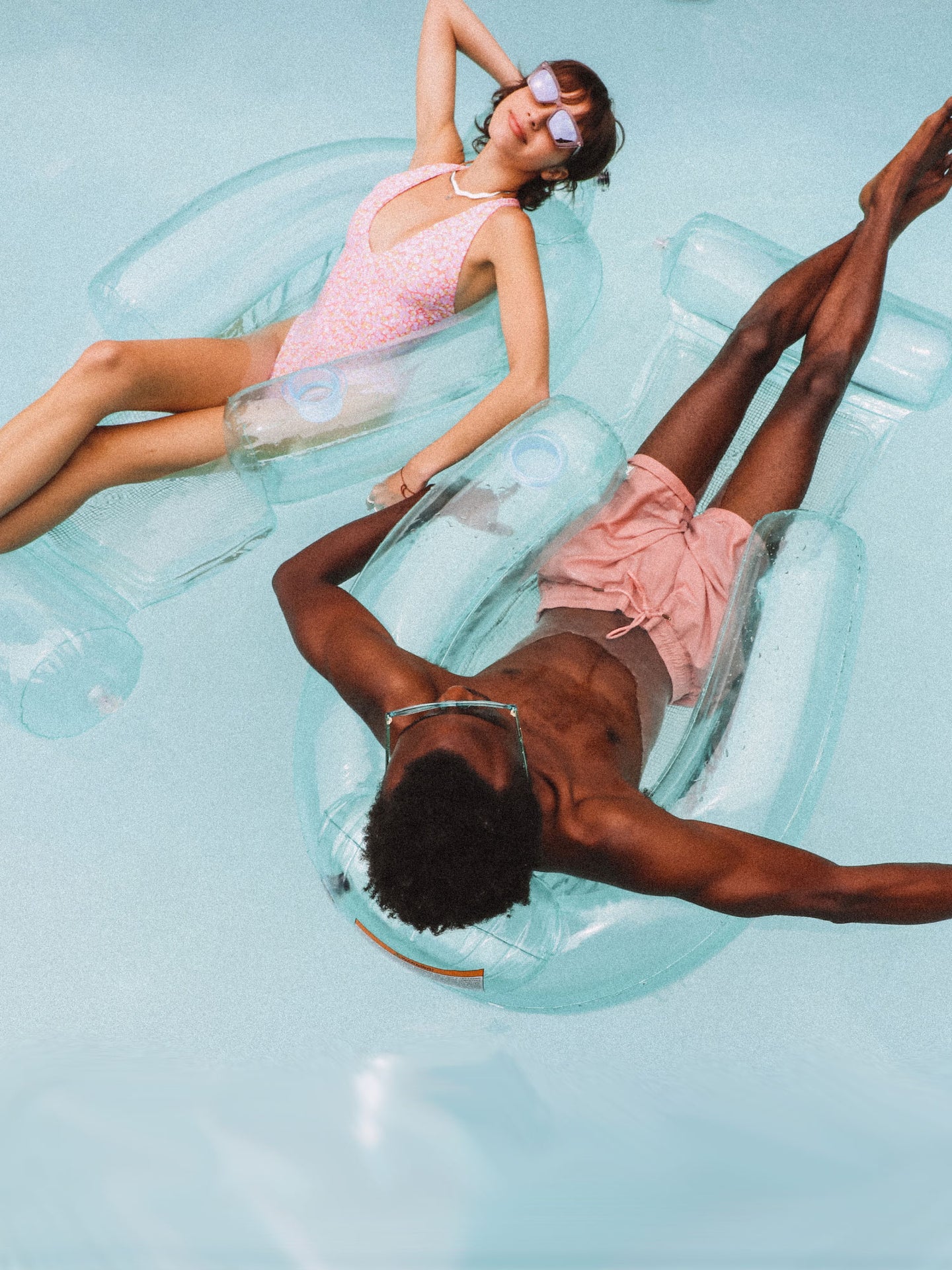 Two friends are each floating on a FUNBOY Clear Blue Mesh Chair in a blue swimming pool. It's a top down shot of the pool.
