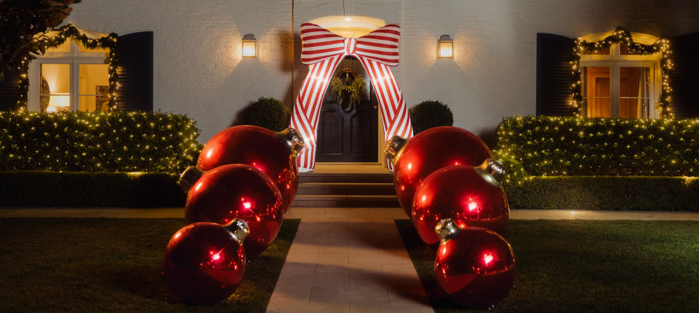 A festive holiday front yard at night with a pathway leading to a decorated house entrance. Large red ornaments line both sides of the walkway, and a red-and-white striped arch with a glowing light fixture is centered above the front door.