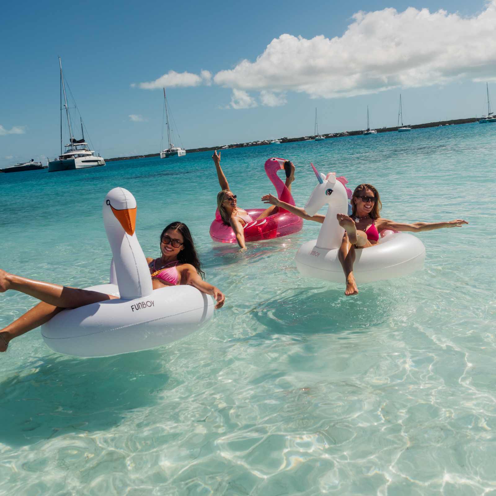 Three women on inflatable pool toys in clear blue water with sailboats in the background.