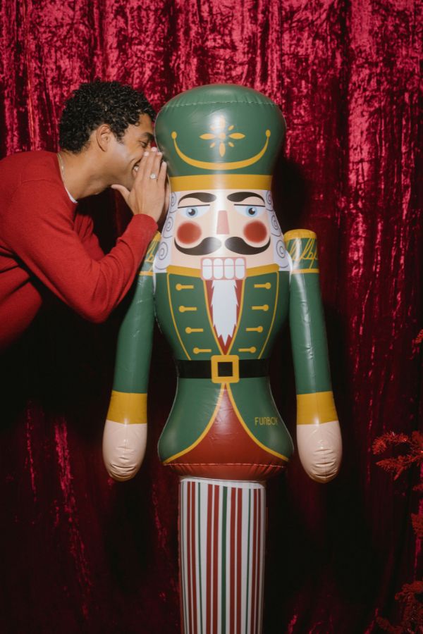 A man in a red sweater whispers into the ear of a large inflatable nutcracker figure with a green and gold uniform, posed in front of a red velvet curtain.