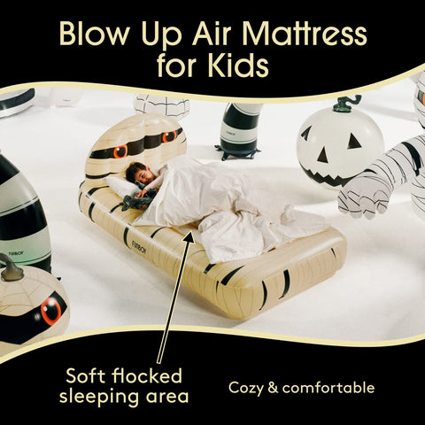 An inflatable mummy-themed air mattress for kids is shown with a child lying underneath and white bedding. The scene includes Halloween props like a jack-o'-lantern and a mummy bandage, with text reading 'Blow Up Air Mattress for Kids' and 'Soft flocked sleeping area.
