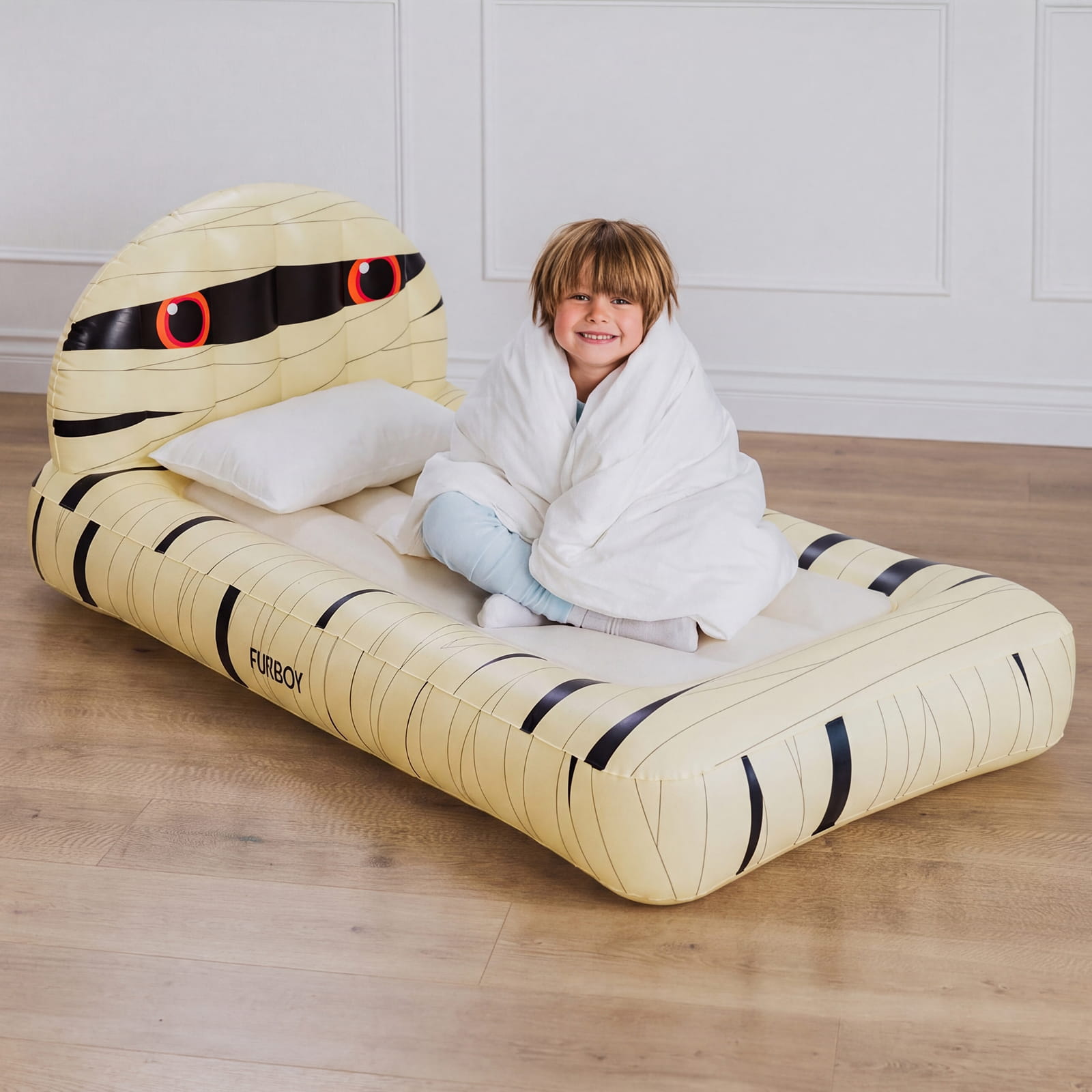 A child sitting on a large inflatable mummy bed in a bright room with light wood flooring and white paneled walls. The bed is pale yellow with black striping and red-and-black eye details, and the child is wrapped in a white blanket.