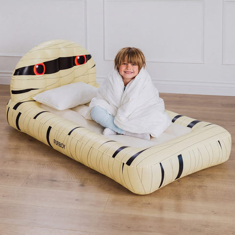 A child sitting on a large inflatable mummy bed in a bright room with light wood flooring and white paneled walls. The bed is pale yellow with black striping and red-and-black eye details, and the child is wrapped in a white blanket.