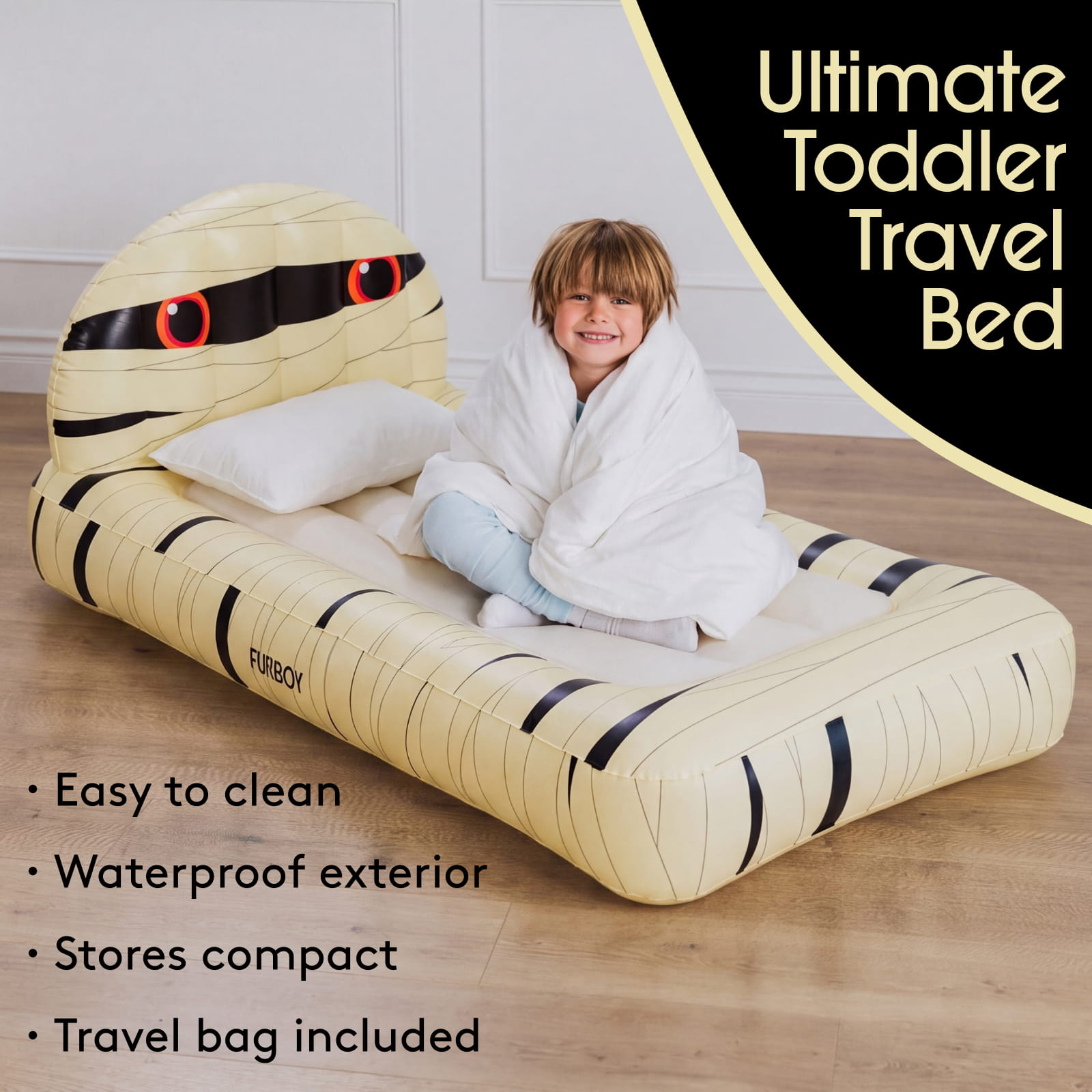 An inflatable toddler travel bed shaped like a cartoon mummy, shown with a child wrapped in a white blanket sitting on it. The bed is cream-colored with black stripes and red eye details.