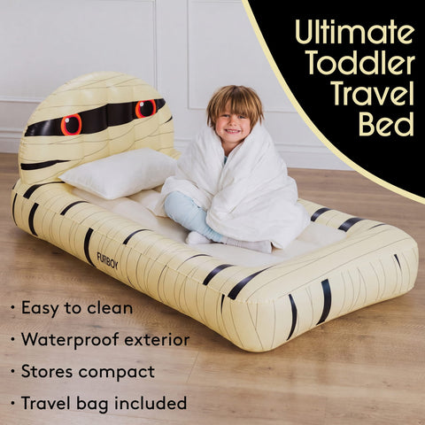 An inflatable toddler travel bed shaped like a cartoon mummy, shown with a child wrapped in a white blanket sitting on it. The bed is cream-colored with black stripes and red eye details.