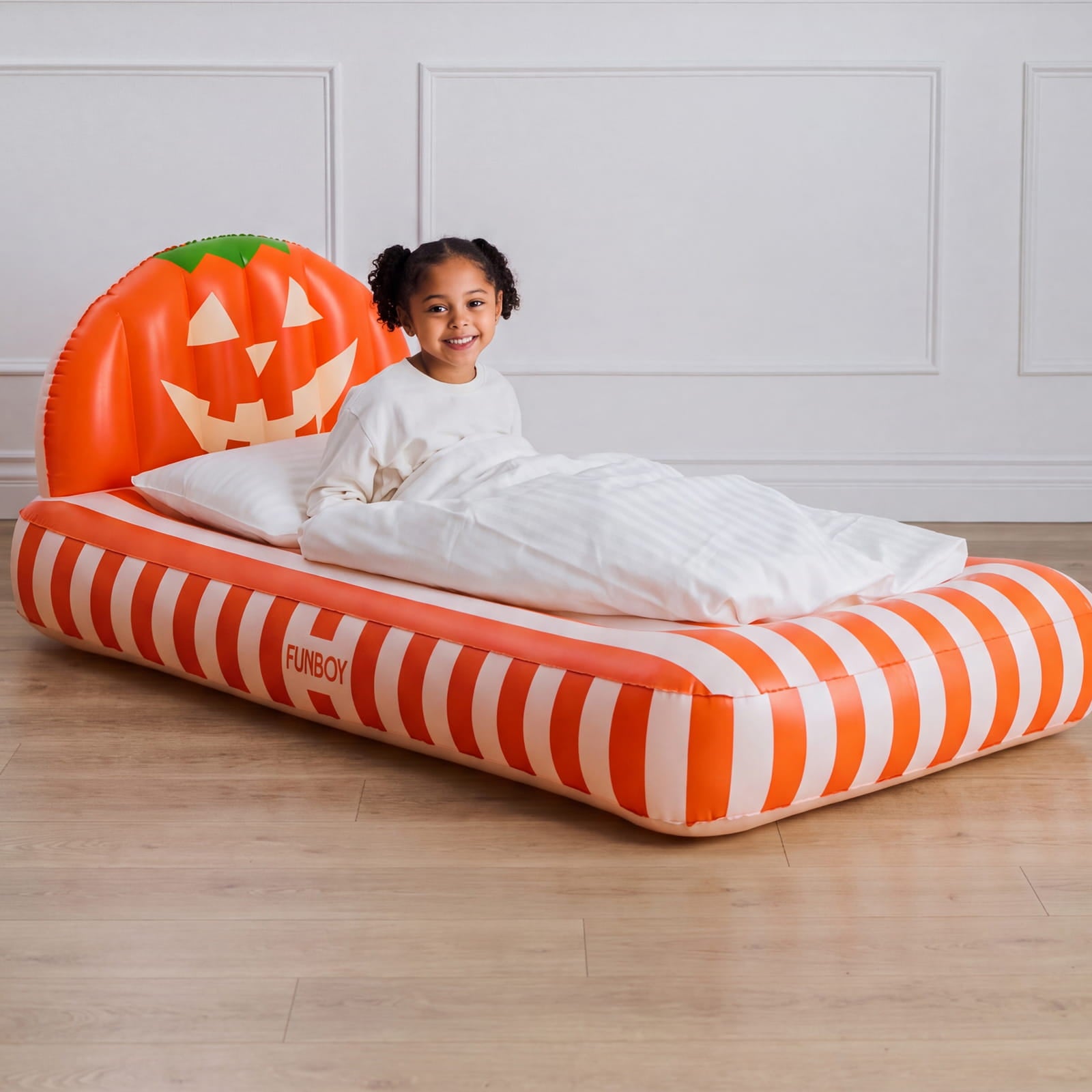 A young girl sits on an inflatable bed with a pumpkin headboard and orange-and-white striped sides, photographed in a bright indoor room with a wood floor and white paneled wall.