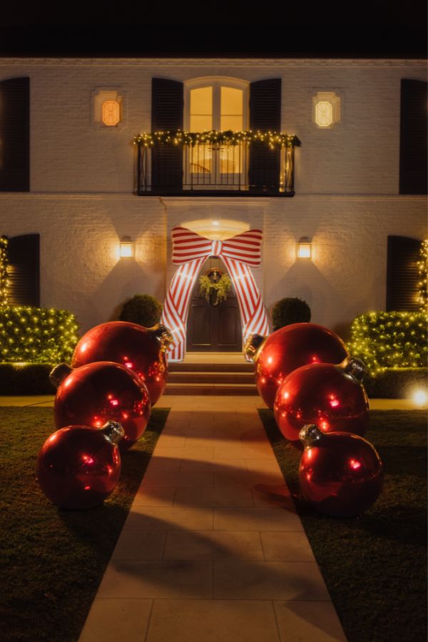 A front yard at night with a house facade, steps, string lights, a large red-and-white striped bow decoration over the door, and several oversized shiny red ornaments placed along the lawn path.