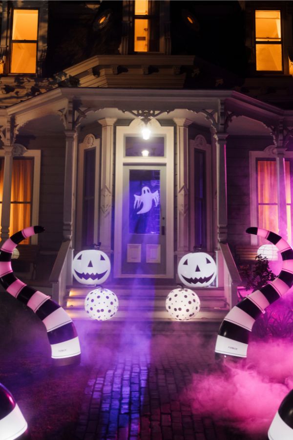 Halloween front yard decoration on a Victorian-style house porch at night, featuring glowing jack-o'-lantern pumpkins, striped black-and-white spiderwebs, and pink and purple fog with a ghost on the front door.