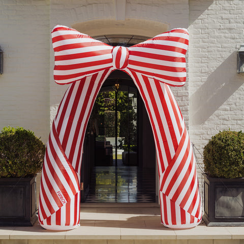 A giant inflatable red and white striped bow arch stands in front of a white brick building entrance, flanked by two potted green shrubs.
