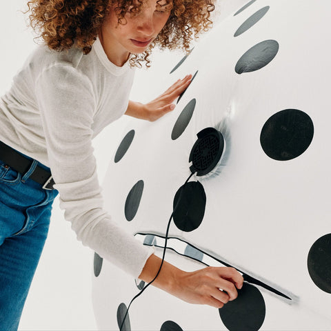 A woman with curly hair wearing a white long-sleeve shirt and blue jeans is shown interacting with a large white surface covered in black polka dots. A black circular port with a mesh cover and a cord is attached to the surface.