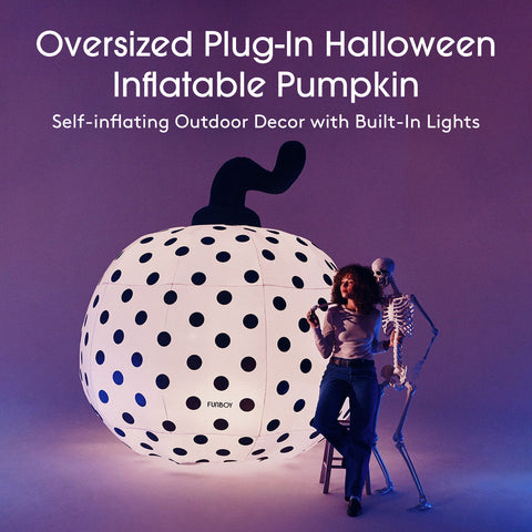 An oversized plug-in Halloween inflatable pumpkin in white with black polka dots and a black stem, shown next to a woman and a small skeleton prop in a studio setting with purple lighting. Text at the top reads: 'Oversized Plug-In Halloween Inflatable Pumpkin' and 'Self-inflating Outdoor Decor with Built-In Lights.