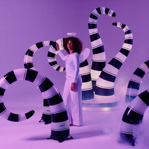 A woman in a white angel outfit stands among large black-and-white striped inflatable serpent-like props in a purple-lit studio setting.