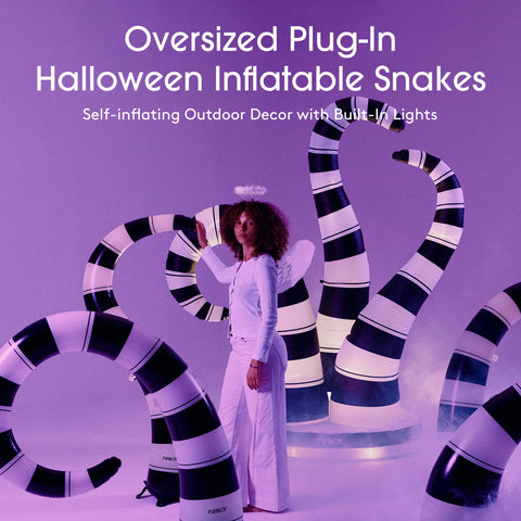 A person stands in a studio wearing a white outfit and posing with oversized inflatable snakes that are glowing with black-and-white stripes. Large text at the top reads 'Oversized Plug-In Halloween Inflatable Snakes' and 'Self-inflating Outdoor Decor with Built-In Lights.