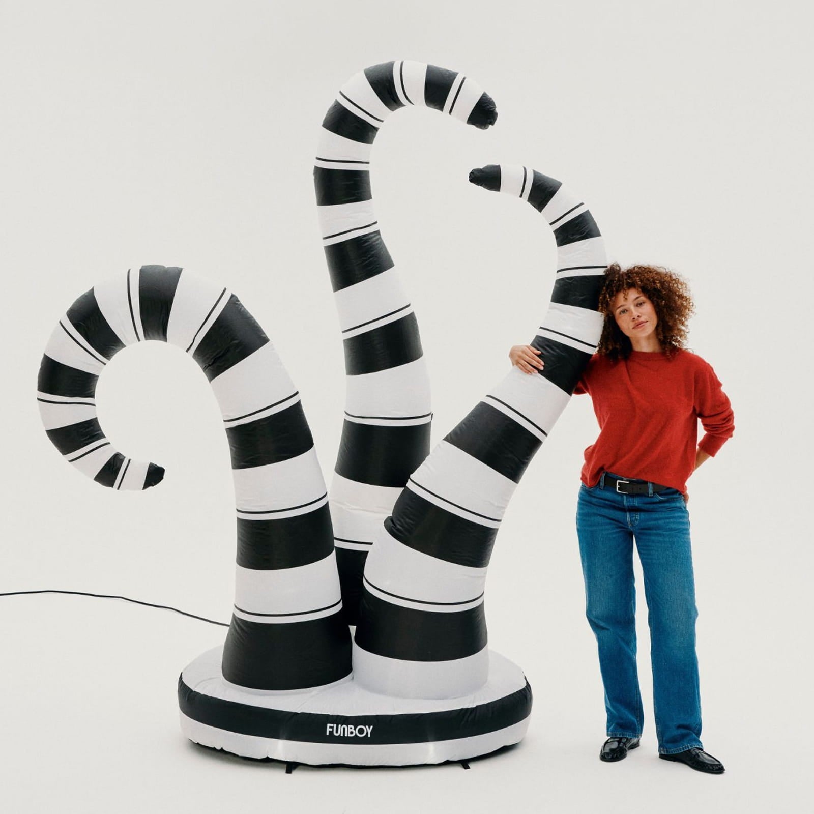 A person stands beside a large inflatable black-and-white striped monster-like sculpture on a white studio background. The product has multiple curved tentacle-like forms rising from a round base, and the base is labeled FURBOY.