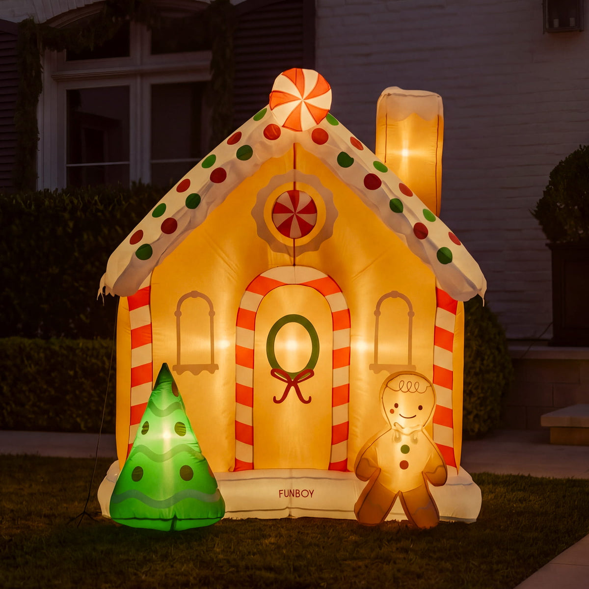 An inflatable gingerbread house holiday decoration lit up at night, displayed on a lawn in front of a house. The decoration features a warm yellow body, red and white striped trim, green and white polka dots on the roof, a candy cane wreath on the door, a green tree, and a gingerbread man figure.