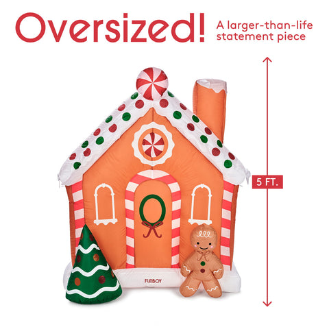 An oversized gingerbread house inflatable statement piece by FURBOY, shown on a white background with red text indicating it is 5 feet tall and larger than life. A small green Christmas tree and an inflatable gingerbread person are placed in front of the house.
