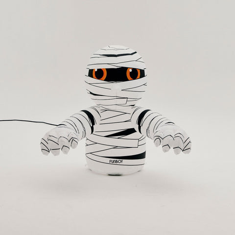 A white and black mummy-themed toy or plush figure with orange eyes, shown against a plain light gray background. A black power cord extends from the left side, and the brand name 'FLYBECH' is printed near the base.
