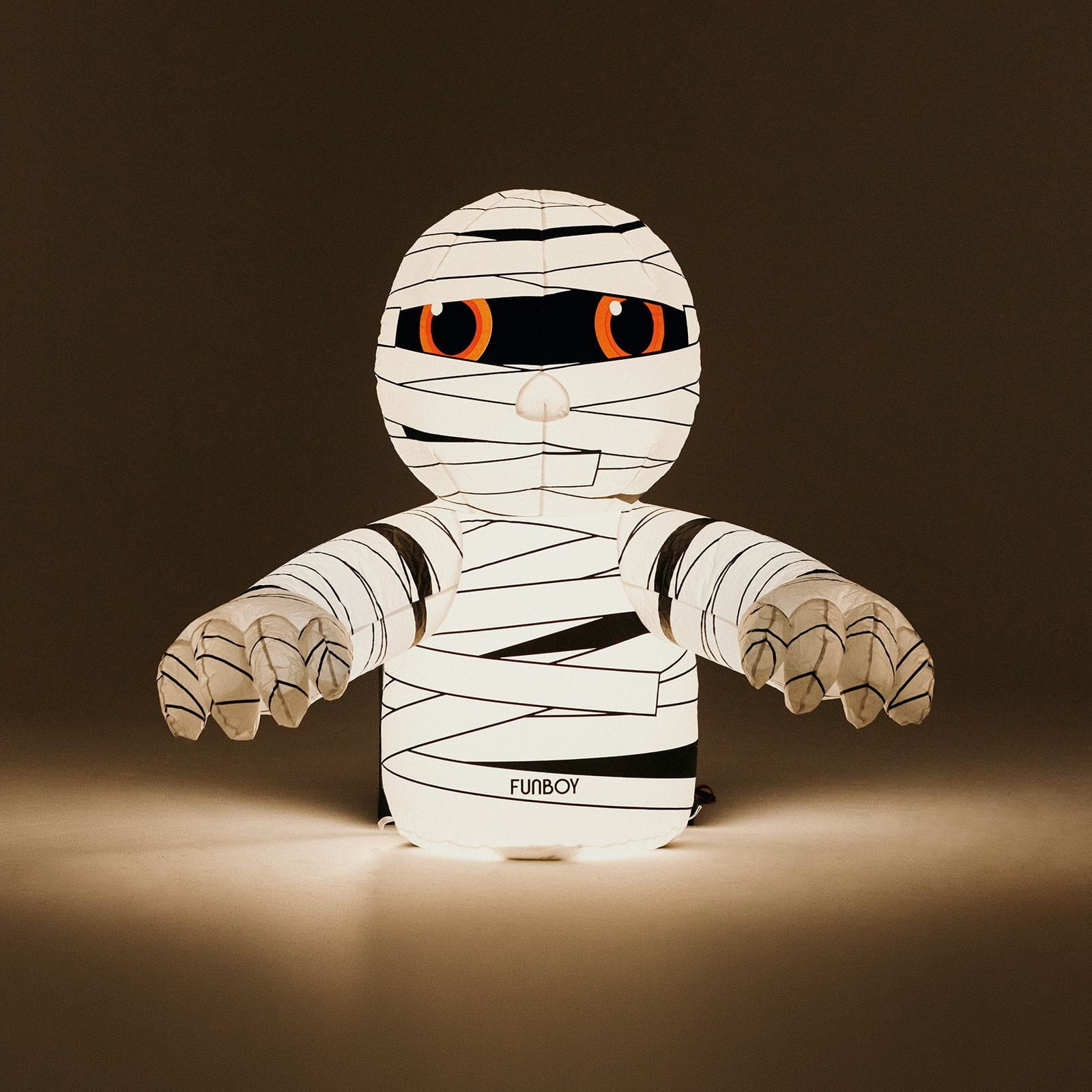 A mummy-shaped LED night light with white bandages, black details, and glowing orange eyes, set against a dark brown background.