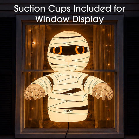 A glowing mummy-shaped window decoration displayed in a window frame, with warm string lights around it and overlaid text reading 'Suction Cups Included for Window Display.' The mummy has white banding, orange eyes, and the name 'FUNBOY' on its torso.