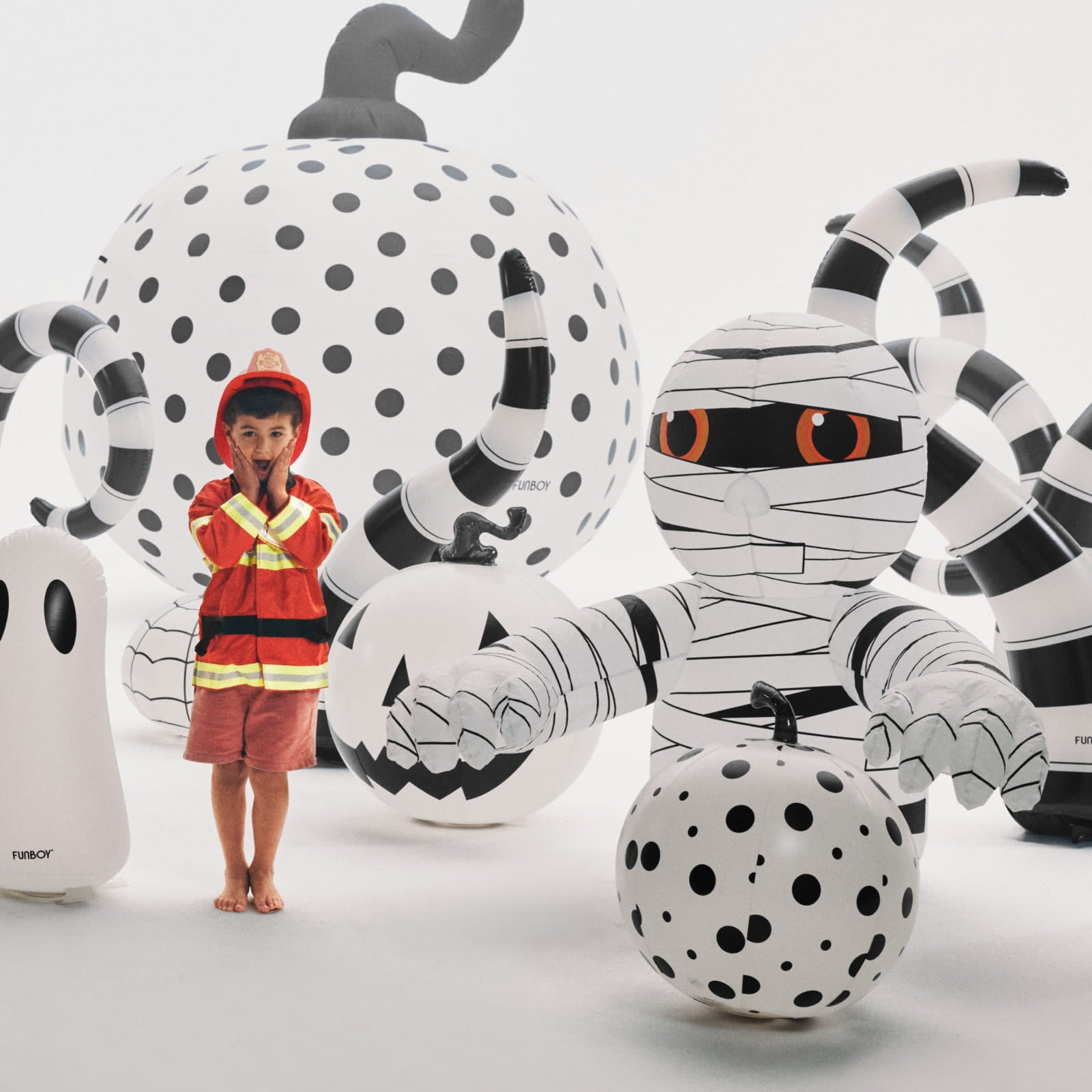 A child in a firefighter costume stands barefoot among large inflatable Halloween decorations, including a mummy, polka-dot pumpkins, and a ghost, in a bright white studio setting.