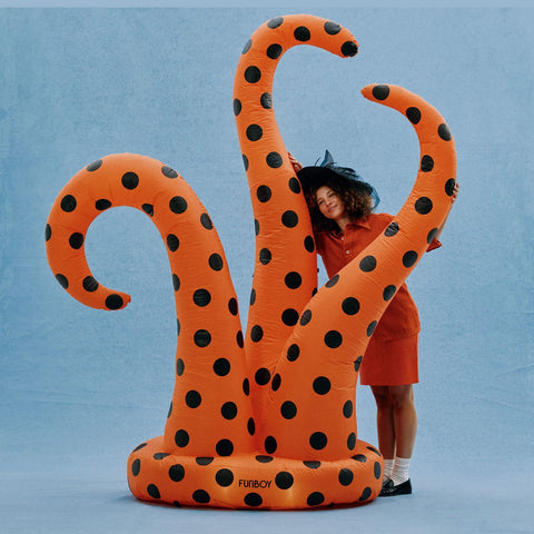 An orange inflatable octopus tentacle with black polka dots, shown with a woman in an orange dress and black witch hat peeking behind it against a light blue studio background.