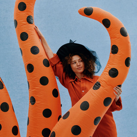 A woman in an orange jumpsuit and black witch hat poses behind a large orange inflatable tent with black polka dots against a light blue background.