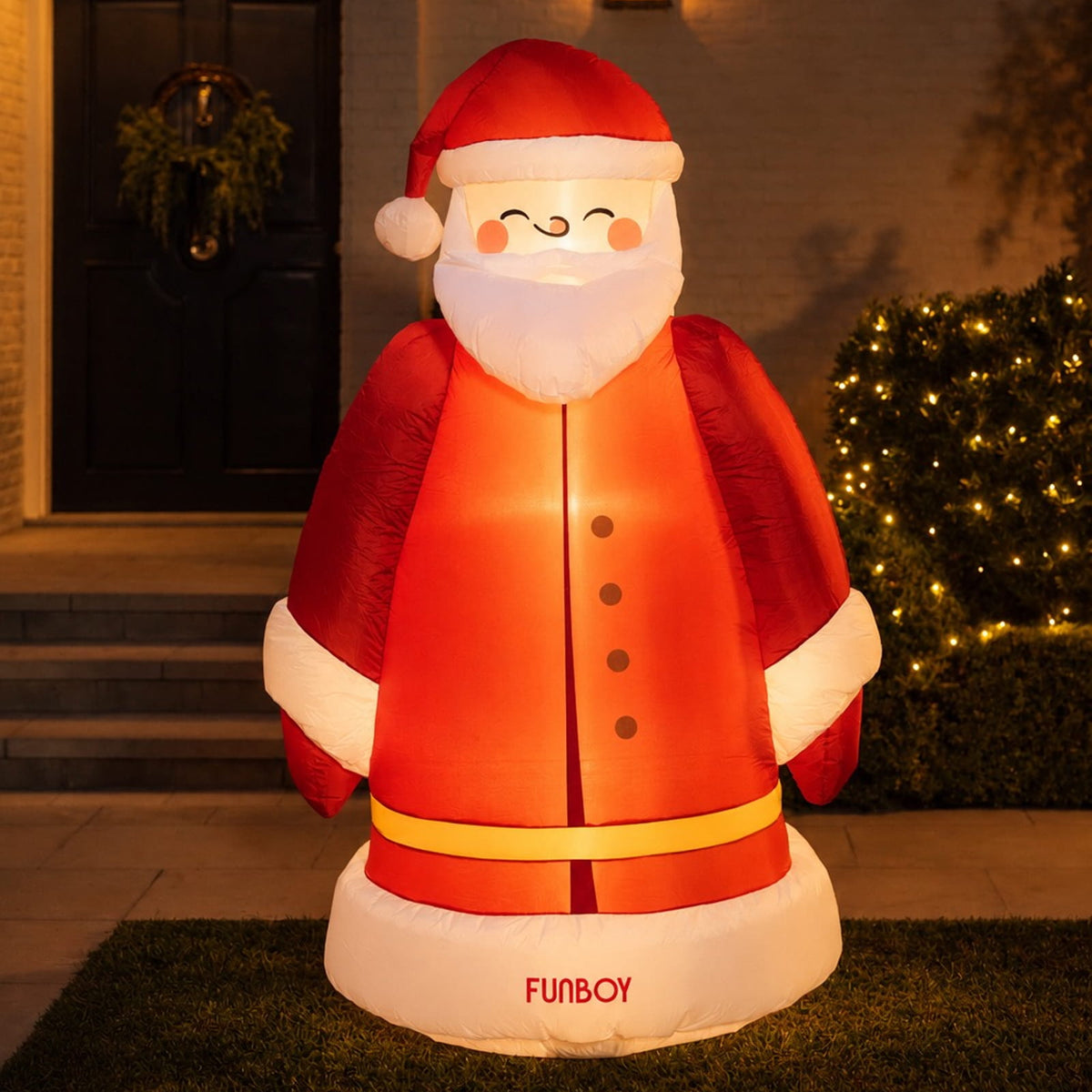 An inflatable Santa Claus figure stands on a lawn in front of a house at night. The glowing Santa is dressed in red with white trim and a yellow belt, and the words "FUNBOY" are printed on the white base.