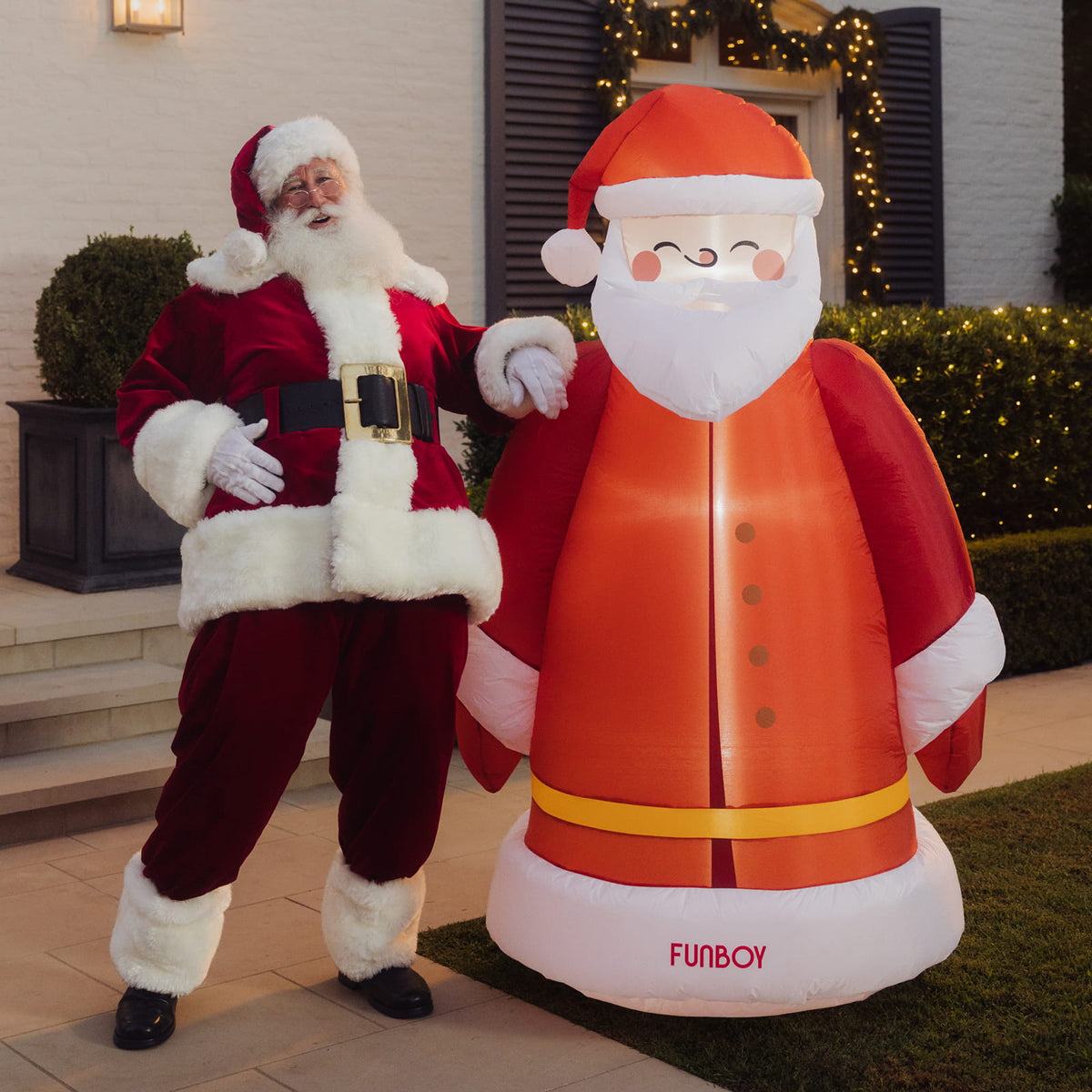 A person dressed as Santa Claus stands beside a large inflatable Santa-shaped lighted toy outdoors in front of a house with lit holiday garlands. The inflatable is red and white with a yellow belt and the word 'FUNBOY' near the base.