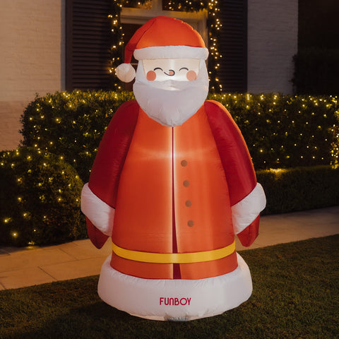 An inflatable Santa Claus figure stands on a lawn in front of a house at night, with lit bushes and a window in the background. The figure is mostly red and white with a yellow belt and the word "FUNBOY" printed near the base.