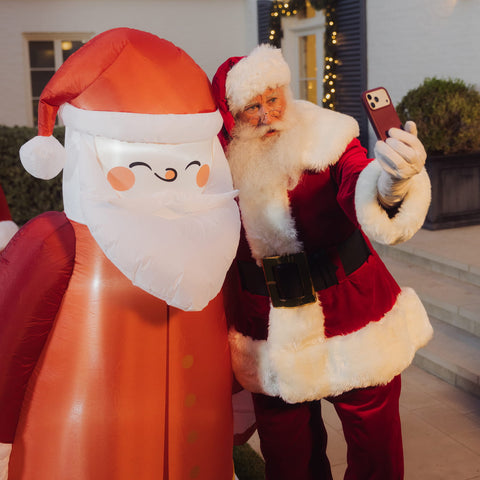 A person dressed as Santa Claus taking a selfie next to a large inflatable Santa decoration in front of a house exterior.