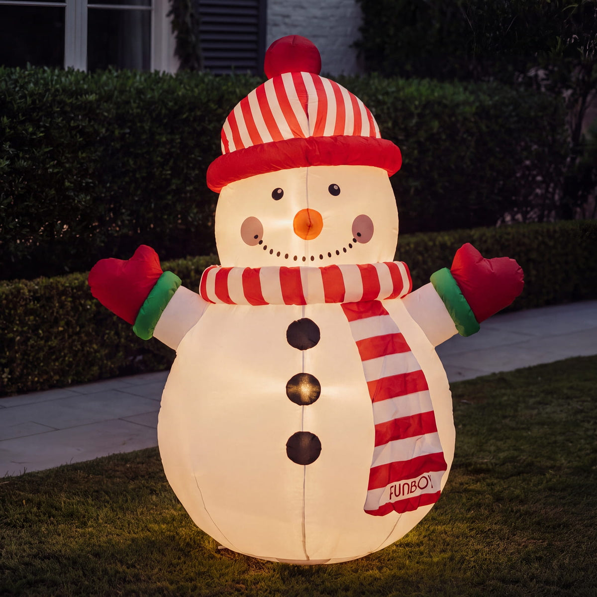 A large inflatable snowman light standing on grass in front of a hedge at night. The snowman is glowing from within, wearing a red-and-white striped hat, scarf, and gloves, with a red nose and black buttons.