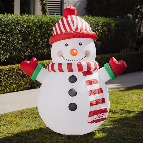An inflatable snowman decoration standing on a grassy lawn in front of trimmed hedges. The snowman is white with a red-and-white striped hat, matching striped scarf, red mittens, green cuffs, black buttons, and an orange nose.
