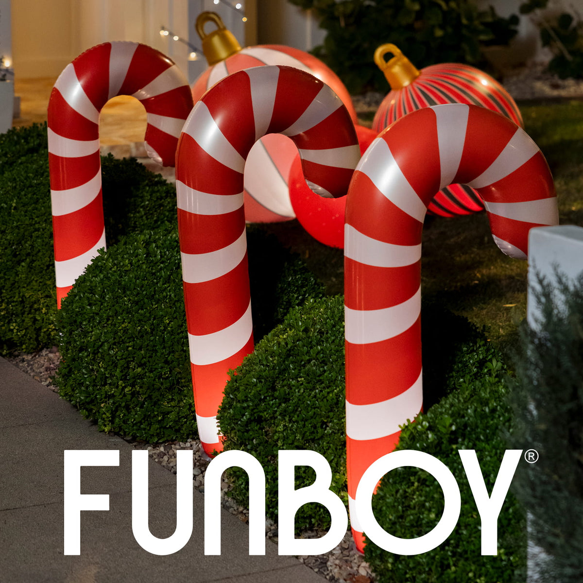 Large inflatable holiday decorations shaped like candy canes and ornaments displayed outdoors in front of trimmed green hedges at night, with the FUNBOY logo overlaid across the bottom.