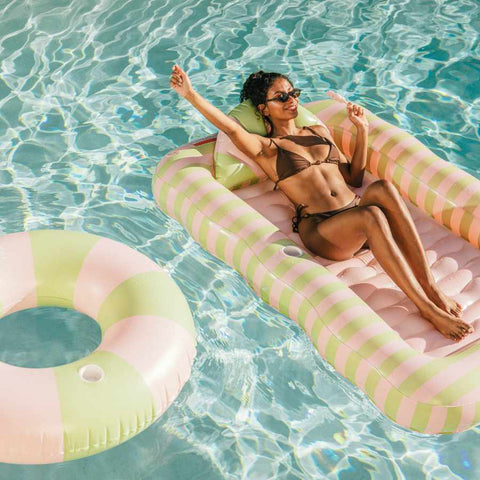 Woman relaxing on a striped inflatable pool float tanning lounger in a pool with a pink and green inner tube nearby.