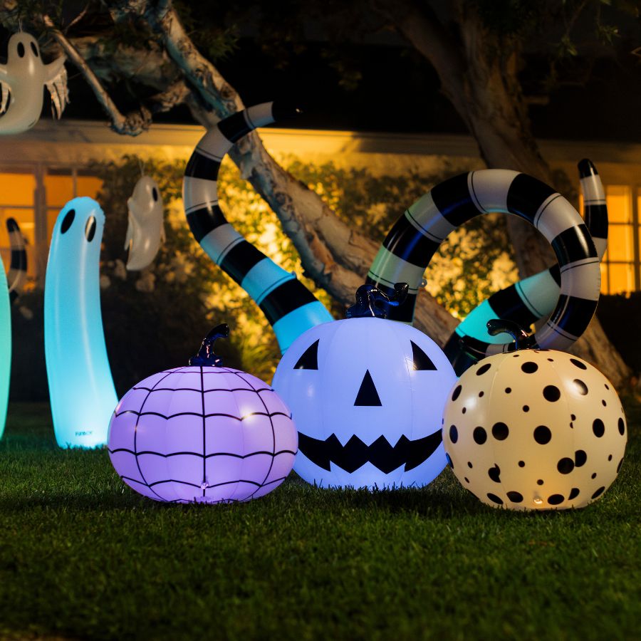 Inflatable Halloween yard decorations on grass at night, including a purple spiderweb pumpkin, a glowing blue jack-o'-lantern, a cream polka-dot pumpkin, a striped snake, and ghost figures.