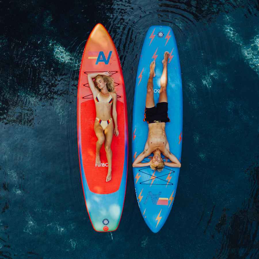 Two people lying on paddleboards in the water, one red and one blue.
