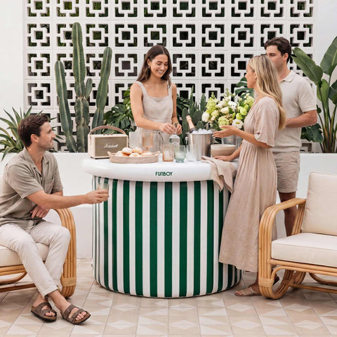 Group of people gathered around a green and white striped bar in an outdoor setting with plants and a geometric wall design.