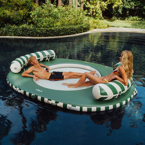 Two people relaxing on a green and white striped inflatable pool float in a pool surrounded by greenery.