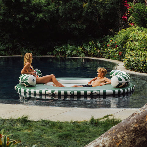 Two people on a green and white inflatable pool toy in a pool surrounded by greenery.
