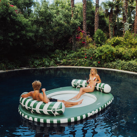 Two people on green and white striped pool floats in a pool surrounded by lush greenery.