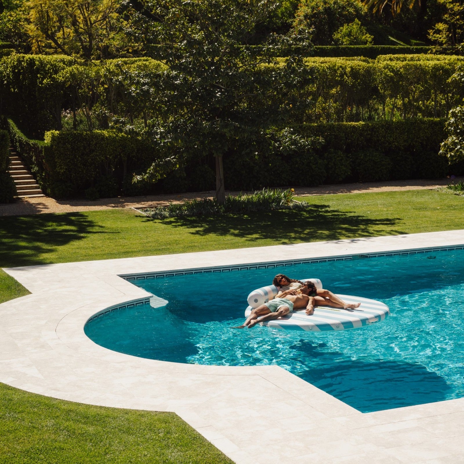 Two people on a inflatable pool float in a swimming pool surrounded by greenery.