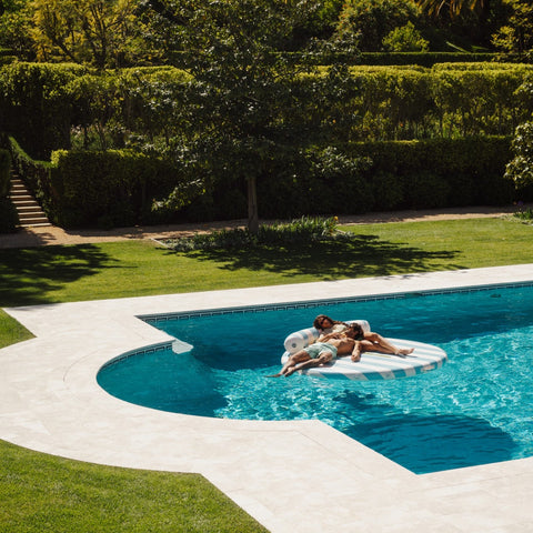 Two people on a inflatable pool float in a swimming pool surrounded by greenery.