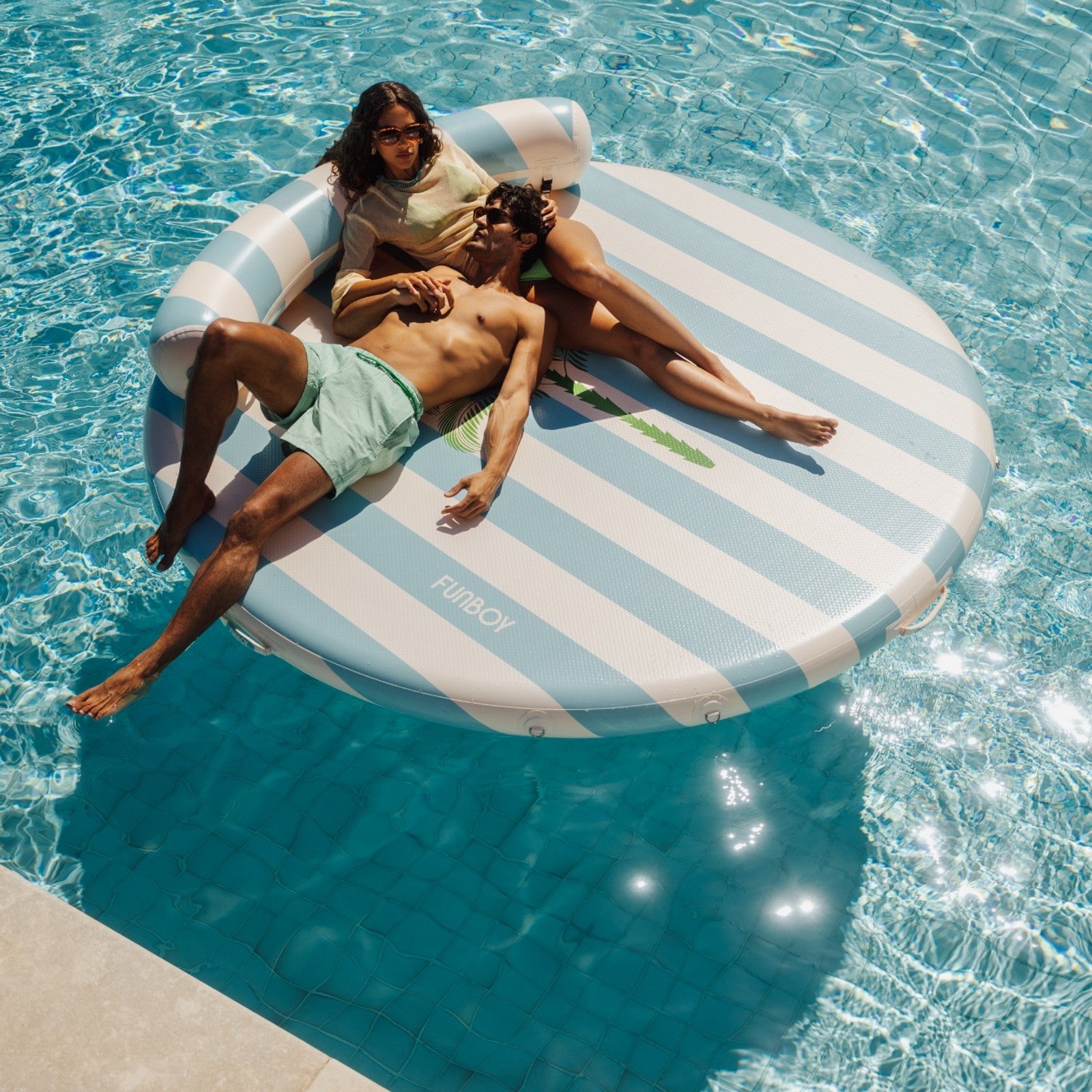 Two people lounging on a striped inflatable pool float in clear blue water.
