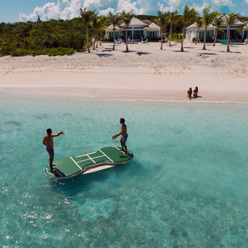 Two people playing tennis on a floating pickle bar inflatable in clear blue water with a beach and palm trees in the background.