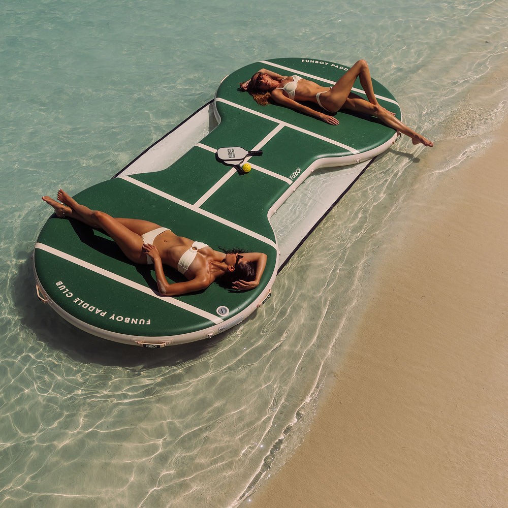 Two women lying on a paddle ball court float in clear water near a sandy beach.