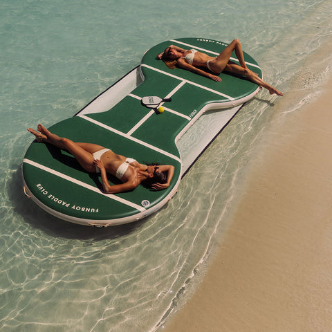Two women lying on a paddle ball court float in clear water near a sandy beach.