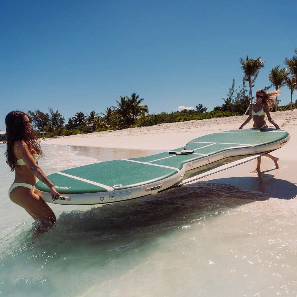 Two women carrying a large green and white inflatable board on a sandy beach with palm trees in the background.