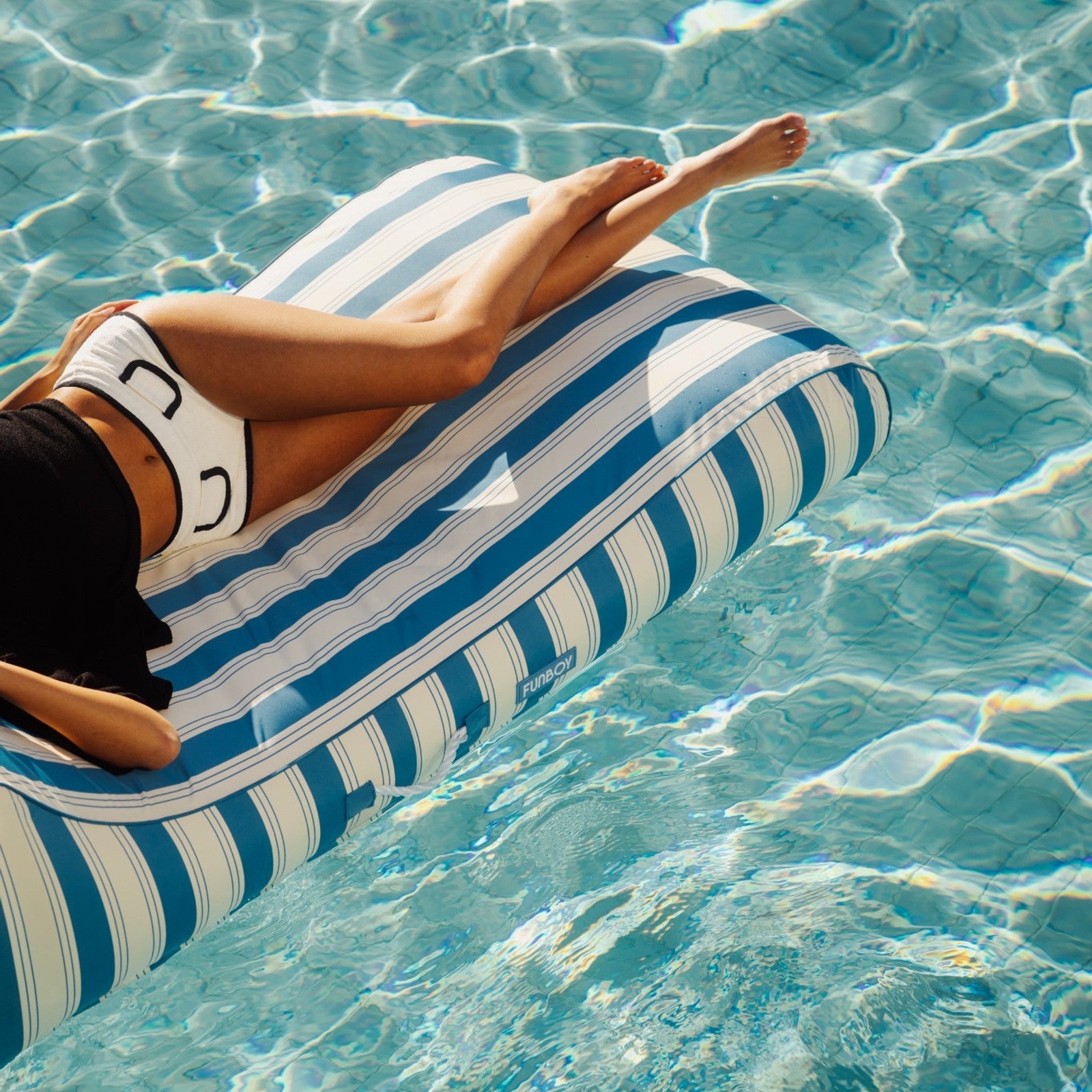 Person lounging on a blue and white striped inflatable pool float in clear water.