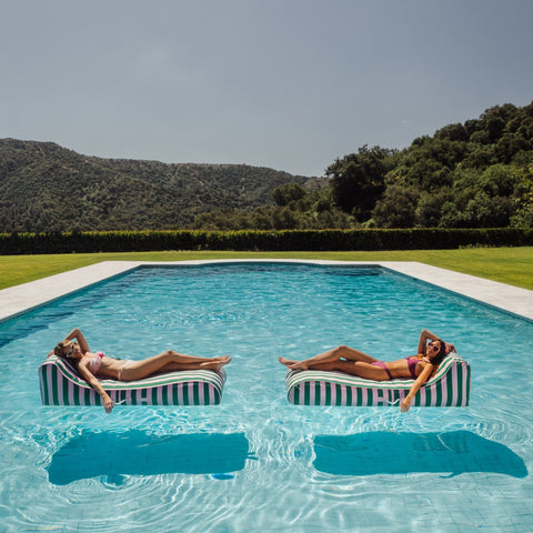 Two women lounging on striped pool floats in a pool with a scenic background.